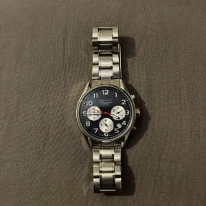 Silverfärgad klocka från GANT - Stilren herrklocka från GANT med silverfärgad länk i metall och svart urtavla. Klockan har tre mindre vita urtavlor, tydliga siffror och röda detaljer på visarna. Perfekt accessoar för dig som gillar klassisk design. Får även med boxen vid köp.