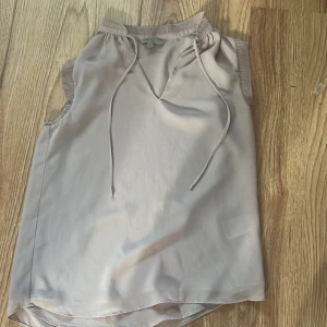 Beige ärmlös topp från H&M - Säljer en stilren beige ärmlös topp från H&M med ribbade detaljer vid hals och ärmhål. Toppen har v-ringning med knytband och är tillverkad i ett mjukt, lätt material. Perfekt för varma dagar eller lager på lager. Pris kan diskuteras