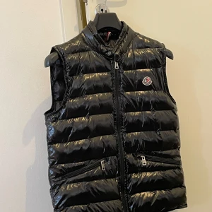 Svart dunväst från Moncler - Snygg svart dunväst från Moncler med glansig finish. Västen har ståkrage, dragkedja framtill och två fickor med dragkedja. Moncler-logga på bröstet. Perfekt för lager-på-lager-stil.