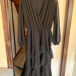 Svart långklänning med volanger från NA-KD - Säljer en svart långklänning från NA-KD med v-ringad omlottfront, långa ärmar i mesh och flera lager volanger på kjolen. Perfekt för dig som vill ha en elegant och stilren look med extra rörelse i kjolen.