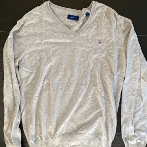 Grå v-ringad tröja från GANT - Säljer en klassisk grå v-ringad tröja från GANT med broderad logga på bröstet. Tröjan har långa ärmar och ribbade muddar vid hals, ärmslut och nederkant. Perfekt för en stilren och enkel look. Tight passform