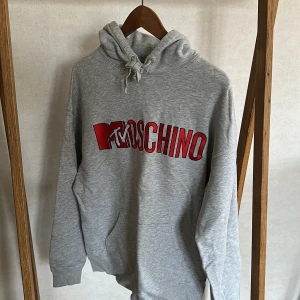Grå hoodie från H&MOSCHINO med röd logga - Säljer en grå hoodie från H&MOSCHINO med stor röd och vit MTVOSCHINO-logga på bröstet.