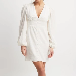 Vit kortklänning - Superfin vit kortklänning från NA-KD med v-ringning och långa puffärmar. Klänningen har små pricka detaljer, knappar framtill och markerad midja. Perfekt för vår och sommar! Har använt en gång💕