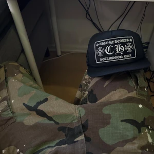 Svart keps från Chrome Hearts - Snygg svart keps från Chrome Hearts med ett stort broderat märke framtill. Märket har vita detaljer och texten 'Chrome Hearts Hollywood, USA'. Kepsen har en klassisk form med justerbar passform och nät på baksidan för ventilation.