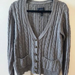 Grå kofta med knappar - Grå kabelstickad kofta från Forever 21 med v-ringning, stora fickor framtill och dekorativa metallknappar. Koftan har långa ärmar och ribbade muddar vid ärmslut och nederkant. Man kan ha den öppen eller stängd. Storlek M men passar som S. Säljer för 50kr 🌸