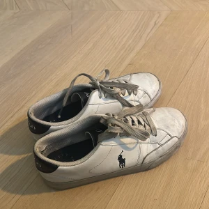 Vita sneakers från Polo Ralph Lauren - Klassiska vita sneakers från Polo Ralph Lauren som har mörkblå märke och detaljer. De är använda, men ser fina ut på utsidan därav priset. Köptes på nk för 1100❤️