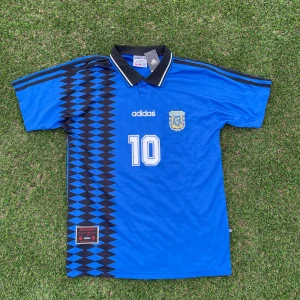 Argentina retro fotbollströja Maradona #10 - Klassisk blå Argentina fotbollströja från Adidas med nummer 10 och Maradona på ryggen. Tröjan har svart och blått mönster längs ena sidan, vit Adidas-logga och landslagsemblem på bröstet. Pikékrage och korta ärmar.