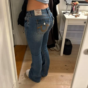 Blå bootcut jeans från True Religion - Säljer ett par klassiska blå jeans från True Religion med bootcut-passform. Jeansen har snygga kontrastsömmar, broderade bakfickor med lock och knappar samt en tydlig logga på bakre linningen. Perfekta för dig som gillar en tidlös och cool denimstil. Storlek 28
