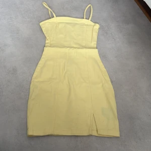 Ljusgul kort klänning med smala axelband - Säljer en ljusgul kort klänning med smala axelband och figurnära passform. Klänningen har en snygg slits framtill och dragkedja i ryggen för enkel påklädning. Perfekt för sommarens alla tillfällen!