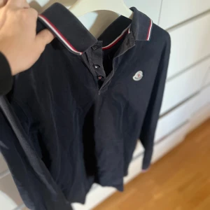 Mörkblå pikétröja från Moncler - Säljer en mörkblå långärmad pikétröja från Moncler med klassisk krage och knappar framtill. 