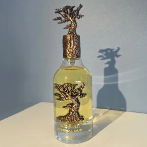 Eternal Oud parfym från Lattafa - Unik parfymflaska med guldfärgad vätska och dekorativt bronsfärgat träd som lock och detalj på flaskan. Flaskan är genomskinlig och har texten 'Eternal Oud' i guld. Perfekt för dig som gillar exklusiva och konstnärliga doftflaskor.