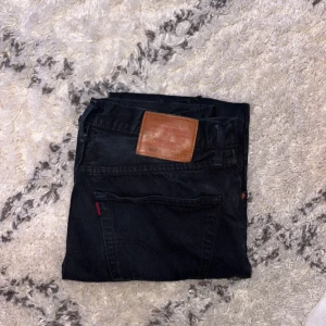 Svarta jeansbyxor Levis 501 - Klassiska svarta Levis 501 jeans med raka ben och femficksmodell. Byxorna har en vit/grå ton på den svarta grundfärgen med en brun läderpatch bak i midjan och röda Levis-detaljer vid bakfickan. Perfekta för en stilren och tidlös look.