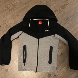Nike hoodie med dragkedja i svart och grå - Snygg hoodie från Nike i svart och grå med hel dragkedja och huva. Hoddien har två fickor med dragkedja framtill och vit Nike-logga på bröstet. Perfekt för träning eller chill.
