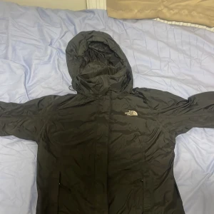 Svart vindjacka från The North Face - Säljer en svart vindjacka från The North Face med huva och dragkedja. Jackan har meshfoder på insidan och klassisk logga på bröstet. Perfekt för blåsiga dagar och enkel att packa ner i väskan.