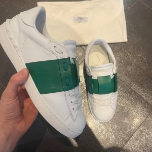 Valentino Open sneakers med grön detalj - Säljer ett par vita Valentino Open sneakers med bred grön läderdetalj över mitten. Skorna har klassisk snörning, rund tå och platt sula. Märkeslogga syns på plösen och insidan. Perfekta för dig som gillar stilrena och exklusiva sneakers. Stolek 41 men passar 41-42. Jätte fint skick. Perfekt till sommaren!!