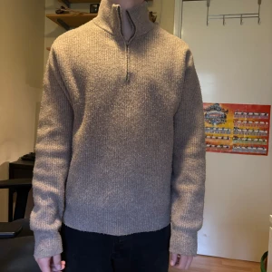 Beige stickad tröja med halv dragkedja - Säljer en beige stickad tröja med hög krage och halv dragkedja. Tröjan har lång ärm och ribbade muddar vid ärmslut och nederkant. Perfekt för kyligare dagar och enkel att matcha med olika outfits.