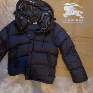 Burberry jacka - Säljer en svart dunjacka från Burberry med avtagbar huva och rutigt innerfoder. Jackan har dragkedja och knappar framtill samt hög krage. Perfekt för kyliga dagar och riktigt snygg design.