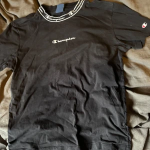 Svart t-shirt från Champion med logga - Svart t-shirt från Champion med vit broderad logga på bröstet och tryckt logga på kragen. Klassisk passform och korta ärmar. Liten Champion-logga även på ärmen. Perfekt för en sportig och avslappnad stil.