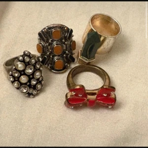 Fyra unika ringar med olika detaljer - Säljer fyra coola ringar: en med vita pärlor och stenar, en med orange detaljer, en med röd rosett och en chunky silverfärgad ring. Perfekta för att ge din stil en personlig touch! Alla ringar för priset !! ❤️