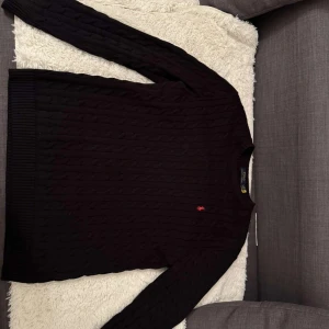 Svart stickad tröja från Ralph Lauren - Snygg svart stickad tröja från Ralph Lauren med kabelstickat mönster och en liten röd logga på bröstet. Perfekt för kyligare dagar med sin klassiska design och långa ärmar.