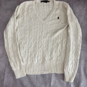 Vit tröja från Ralph Lauren Sport - Kabelstickad vit tröja från Ralph Lauren Sport med v-ringning och broderad logga på bröstet. Tröjan har långa ärmar och ribbade muddar vid ärmslut och nederkant. Klassisk och tidlös design.
