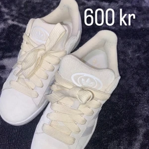 Campus 00s  - Säljer ett par klassiska vita Adidas sneakers med tre vita ränder på sidan och beige gummisula. Skorna har rund tå, platt sula och snörning. Perfekta för en clean och stilren look.