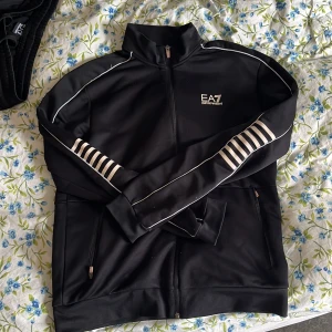 Svarta mjukisbyxor från EA7 Emporio Armani - Svarta mjukisbyxor från EA7 Emporio Armani med vita ränder längs benen och logga på vänster höft. Byxorna har dragsko i midjan och smal passform. Perfekta för en sportig och avslappnad stil. Dessutom får man med en svart tröja som passar perfekt med byxorna 