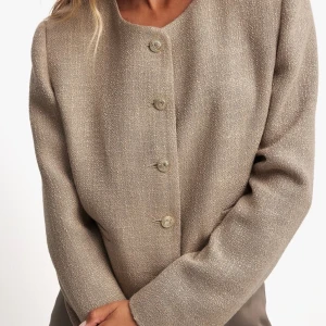 Beige kavaj från NA-KD - Stilren beige kavaj från NA-KD med rund halsringning och raka axlar. Jackan är croppad och har långa ärmar samt knäppning framtill med fem knappar. Passar perfekt till en dressad look och är tillverkad i ett strukturerat tyg. Ny med lapp kvar.