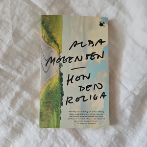 Hon den roliga - Säljer boken 'Hon den roliga' av Alba Mogensen. Omslaget har en landsväg och gröna fält mot en blå himmel, med titeln och författarens namn i svart handskriven stil. Romanen handlar om identitet, vänskap och att hitta sin plats i livet.