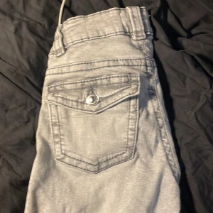Ljusgrå bootcut från gina tricot  - Säljer ett par ljusgrå jeansbyxor från gina tricot. Byxorna har klassisk femficksdesign, bälteshällor och knappgylf. Snygga sömmar och en bakficka med lock och knappdetalj. Perfekta för en stilren vardagslook.
