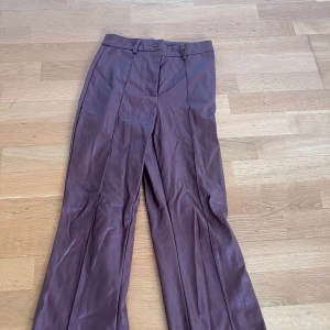 Bruna skinnbyxor med vida ben - Säljer ett par bruna skinnbyxor med vida ben och pressveck. Byxorna har hög midja, bälteshällor och stängs med dragkedja och knapp. Perfekta för dig som vill sticka ut med en cool look.