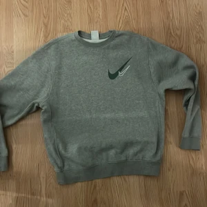Grå sweatshirt från Nike - Grå sweatshirt från Nike med rund halsringning och grönt Nike-logga tryckt på bröstet. Klassisk modell med ribbade muddar vid ärmslut och nederkant. Perfekt för en avslappnad stil.