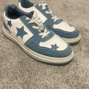 Blåvita sneakers med stjärndetaljer från SRE Sport - Säljer ett par snygga sneakers i vitt och ljusblått från SRE Sport. Skorna har tydliga stjärndetaljer på sidorna och hälen, samt vita snören och en vit sula. Materialet är en mix av syntet och jeansliknande tyg.