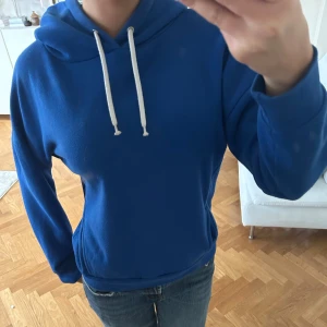 Blå hoodie med vita snören - Säljer en klassisk blå hoodie med huva och vita dragsnören. Tröjan har en normal passform och är tillverkad i ett mjukt material som känns skönt mot huden. Perfekt för en avslappnad stil.