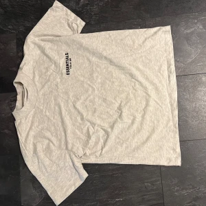 Grå Essentials Fear of God Herr t-shirt  - Snygg ljusgrå t-shirt från Essentials Fear of God med svart tryck både fram och bak. Klassisk passform med rund hals och korta ärmar. Medium men stor i storleken.