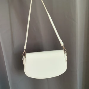 Vit axelväska från Gina Tricot - Stilren vit axelväska från Gina Tricot i syntetmaterial med silverfärgade metalldetaljer. Väskan har en avlång, rundad form och stängs med magnetknapp. Perfekt storlek för det viktigaste och enkel att matcha till olika outfits.