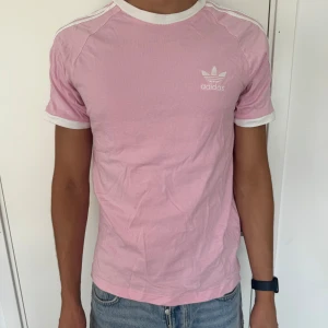 Rosa Adidas t-shirt med vita detaljer - Snygg rosa t-shirt från Adidas med klassiska vita ränder på axlarna och vit logga på bröstet. T-shirten har vita muddar vid ärmslut och hals, och är i mjuk bomull. Perfekt för en sportig och avslappnad look.