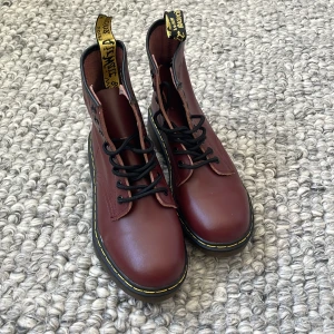 Vinröda Dr. Martens boots i skinn - Säljer ett par klassiska vinröda Dr. Martens boots i skinn med svarta snören och ikonisk gul söm runt sulan. Grov platt sula och rund tå, samt den typiska Dr. Martens-dragfliken baktill, dock är den högra dragfliken trasig men fullt funktionell. Storlek 38. Aldrig använda. 