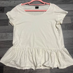 Vit volangtopp från Gina Tricot - Supersöt vit topp från Gina Tricot med volang nertill. T-shirtdesign med rund halsringning och korta ärmar. Perfekt för dig som gillar en enkel men ändå lekfull stil. Tillverkad i mjuk bomull som känns skön mot huden.