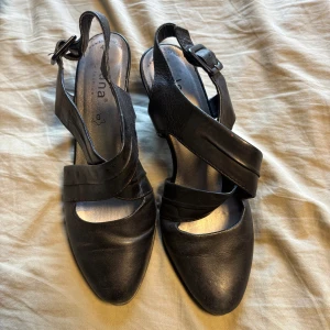 Svarta klackade sandaler från Jana - Snygga svarta sandaler från Jana med klack och korsade remmar i skinn. Skorna har öppen häl, justerbart spänne och spetsig tå. Perfekta för dig som gillar en stilren look med lite edge.
