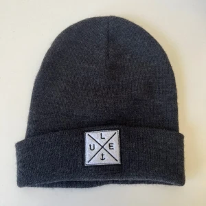 Mörkgrå stickad mössa  - Mörkgrå stickad mössa med uppvikt kant och en vit fyrkantig patch framtill.