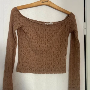 Brun/rosa spetsig offshoulder topp - Säljer en brun/rosa långärmad topp i spets med offshoulder-modell. Toppen har en croppad passform och är tillverkad i ett genombrutet spetsmaterial som ger en cool och trendig look. Perfekt att styla med höga jeans eller kjol.