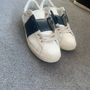 Valentino Garavani Open sneakers vit/blå - Snygga Valentino Garavani Open sneakers i vitt skinn med bred marinblå rem över vristen. Klassisk rund tå, platt sula och coola nitar baktill. Skorna har snörning och guldlogga på plösen. Perfekta för dig som gillar exklusiva och stilrena sneakers.Valentino open  -Storlek 44 -Skick 7.5/10 -Använda i max 3 månader -Inget större slitage än lite färg defekter på framsidan av vänstra skon -Ingen originallåda eller tillbehör medföljer vid köp