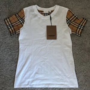 Vit Burberry t-shirt med rutiga ärmar - Vit t-shirt från Burberry med klassiska rutiga korta ärmar i beige, svart och rött. Snygg och stilren design med rund halsringning. Tillverkad i mjuk bomull för en skön känsla. Perfekt för dig som gillar exklusiva streetwear-vibbar. Försöker få sålt snabbt så priset är ej hugget i sten!Aldrig använd!