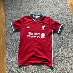 Liverpool röd Nike fotbollströja - Liverpool FCs officiella matchtröja i rött med vita och turkosa detaljer på krage och ärmslut. Tröjan har korta ärmar, klubbmärke och Nike-logga på bröstet samt sponsortryck framtill. Tillverkad i lätt och ventilerande material för maximal komfort.