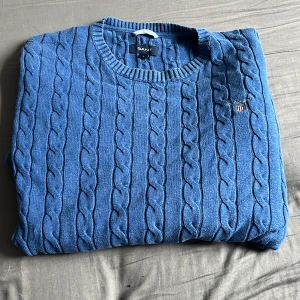 Blå kabelstickad tröja från GANT - Säljer en blå kabelstickad tröja från GANT med rund halsringning och broderad logga på bröstet. Tröjan har klassisk passform och är tillverkad i mjuk bomull, perfekt för lager-på-lager. Snygg till jeans eller chinos! Hör av er vid minsta lilla fråga 