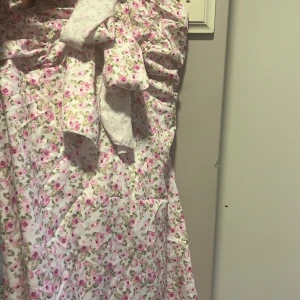 Blommig kortklänning med rosett - Supersöt kortklänning med rosa blommor på vit botten. Finns bättre bilder om man vill ha det i pm.