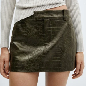 Khakigrön minikjol i skinnlook - Snygg lågmidjad khakigrön minikjol med croco- eller ormskinnsmönster och skinnlook. Kjolen har bälteshällor Helt helt ny oanvänd och med tagg!💚💚