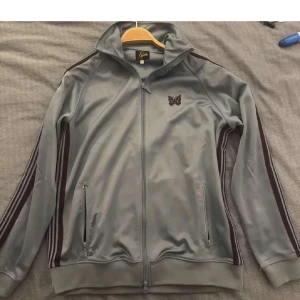 Ljusblå track jacket från Needles L - Säljer en ljus blå track jacket från Needles i storlek L. Jackan har dragkedja framtill, två fickor med dragkedja och mörk lila ränder längs ärmarna. Broderad fjäril på bröstet och klassisk sportig look. Tillverkad i 100% polyester.