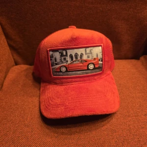 Röd perik keps  - Cool röd keps i sammet från PERIK med en stor patch framtill som visar en sportbil framför ett hus. Kepsen har böjd skärm och klassisk passform, perfekt för dig som gillar bilar och streetstyle.